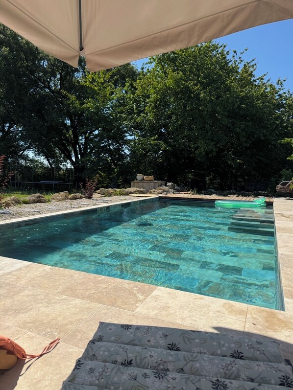 Piscina retangular com água cristalina, rodeada por um deck de pedra e um toldo.