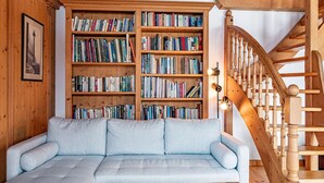 Een gezellige woonkamer met een lichtblauwe modulaire bank, houten boekenplanken vol boeken, een houten trap en een lijst met een foto aan de muur.