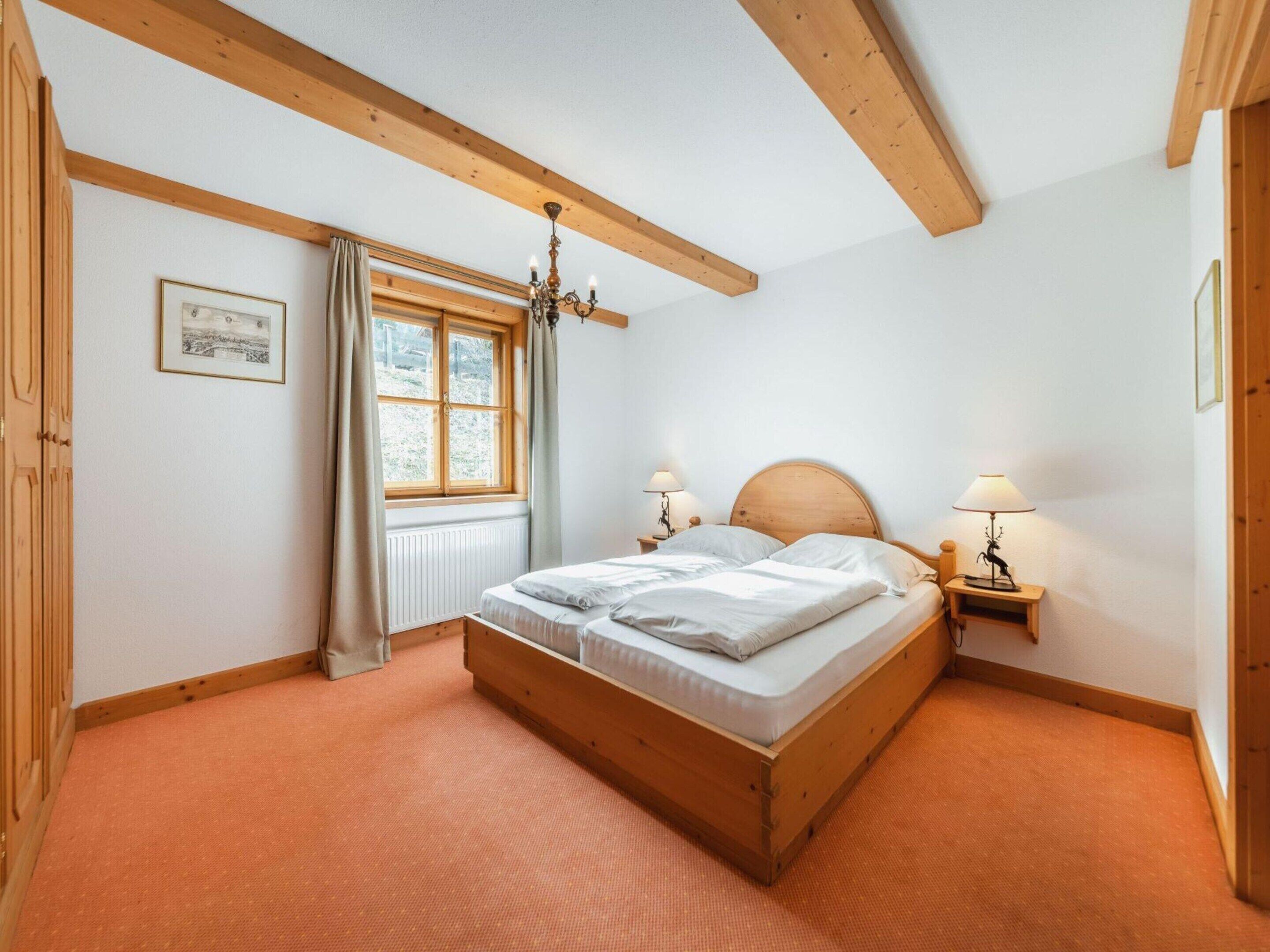 A bedroom with a wooden bed, two bedside tables with lamps, a window with curtains, and a chandelier.