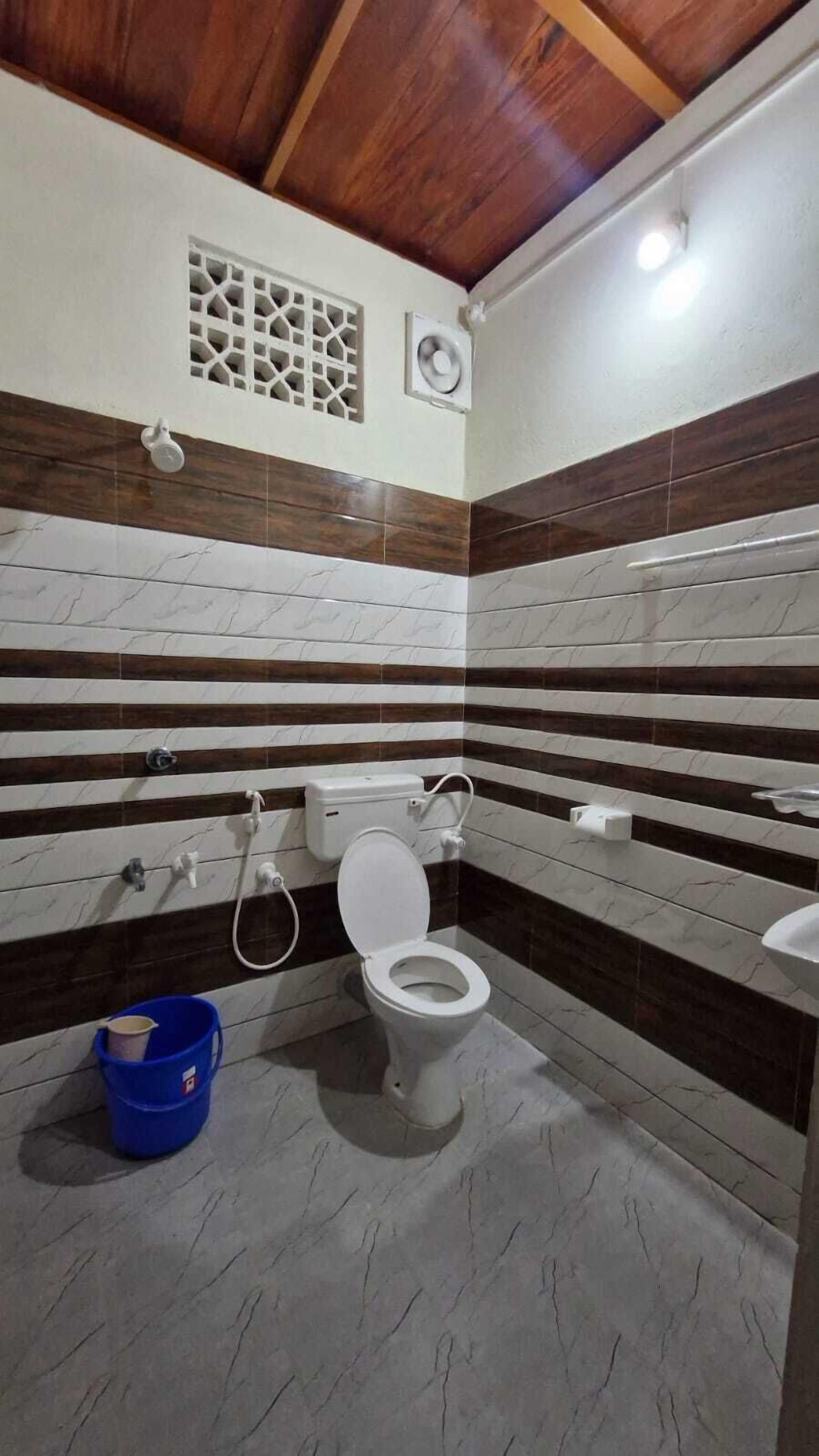 Une salle de bain avec des toilettes, un seau bleu et une bouche d’aération au plafond.
