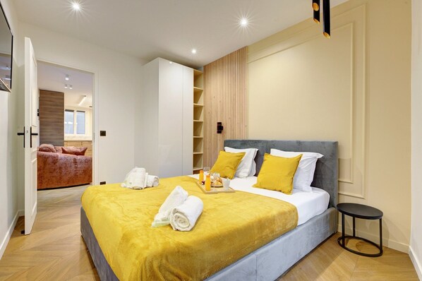 Ein modernes Hotelzimmer mit einem großen Bett, einem Holzschrank, einem kleinen Tisch mit Frühstücksgebäck und Blick auf ein weiteres Zimmer mit einer Couch.
