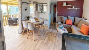 A cozy living area with a wooden floor, a dining table with chairs, a sofa with orange cushions, and a kitchen area with a mirror and shelves. - ZEcamping 4 étoiles familial parc aquatique mobil-home 37m2 (Saint-Jean-de-Monts)