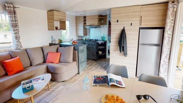 A modern living room with a sofa, a dining table with a magazine and a croissant, and a kitchen area with a refrigerator and a stove. - ZEcamping 4 étoiles familial parc aquatique 100m plage mobil-home clim 36m2 (Canet-en-Roussillon)