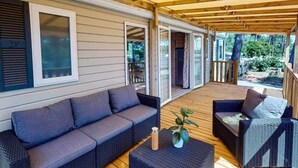 A covered outdoor seating area with a sofa and chairs, wooden deck, and a sliding glass door leading to an interior space. - ZEcamping 4 étoiles animé 2km plage espace aquatique locatif clim 36m2 (La Tremblade)