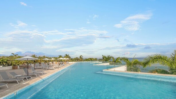 A pool with lounge chairs and umbrellas, surrounded by palm trees and a clear sky.