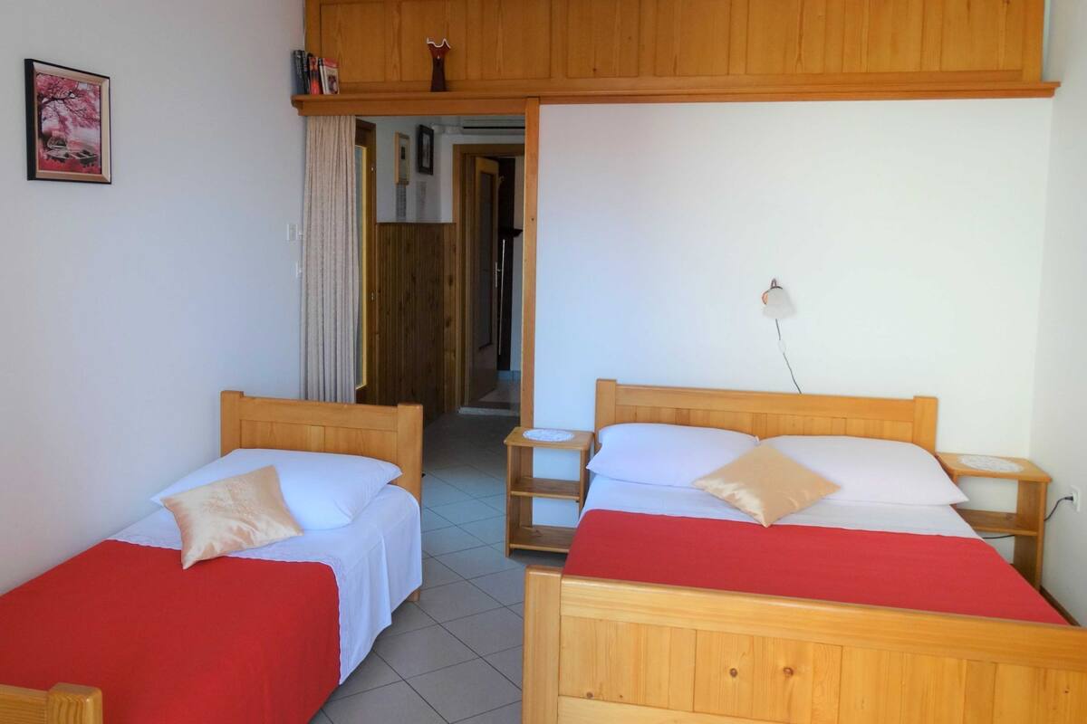 A room with two wooden beds, each with a red bedspread and white linens. There is a wooden cabinet above the beds, a small bedside table with a lamp, and a framed picture on the wall.