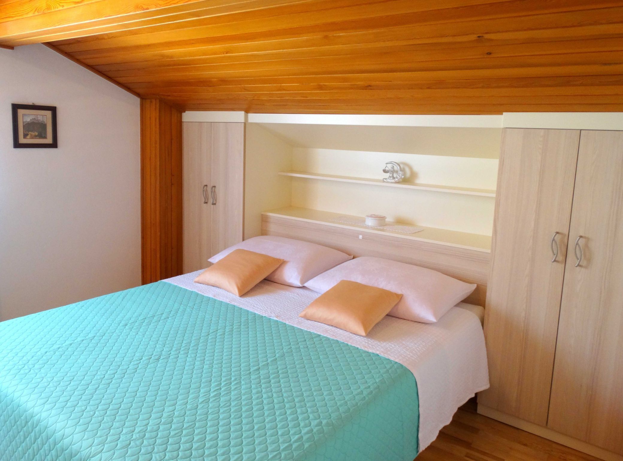 A bedroom with a bed, two pillows, a nightstand, a shelf, and wooden wardrobe.