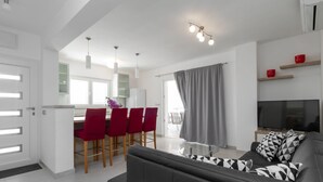 A modern living room with a black leather sofa, a glass coffee table, and a TV on a wooden stand. There is a dining area with red chairs and a white refrigerator. - Modern Villa Near the Sea (Blato)