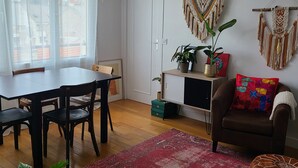 A living room with a dining table, chairs, a red rug, a brown sofa, macramé wall hangings, and a hanging light fixture.