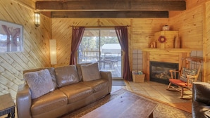 Acogedor interior de cabaña con un sofá de cuero, un sillón mecedor, una chimenea y un ventanal con cortinas.