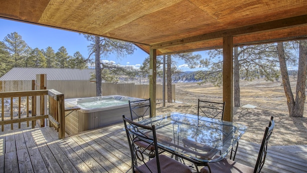 Una terraza de madera con una mesa y sillas de vidrio, un jacuzzi y vistas a árboles y un edificio al fondo.