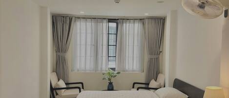A hotel room with two beds, a sitting area with chairs, a window with curtains, a standing fan, and a bedside lamp.