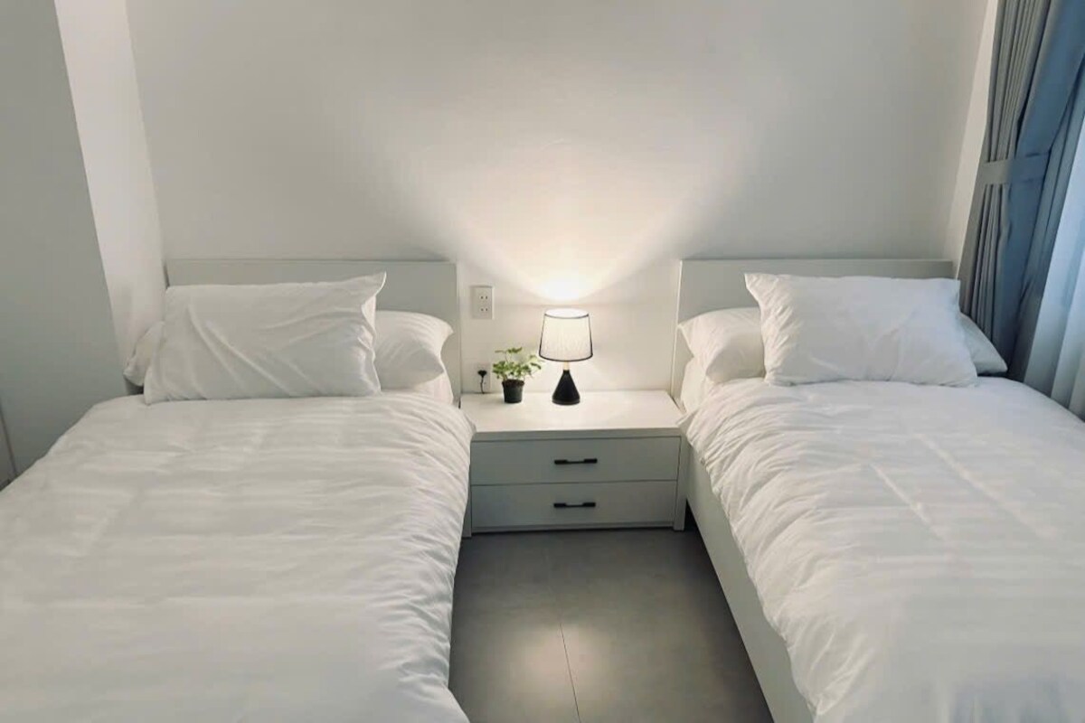 Habitación doble con dos camas, una mesita de noche con lámpara y una pequeña planta en maceta.