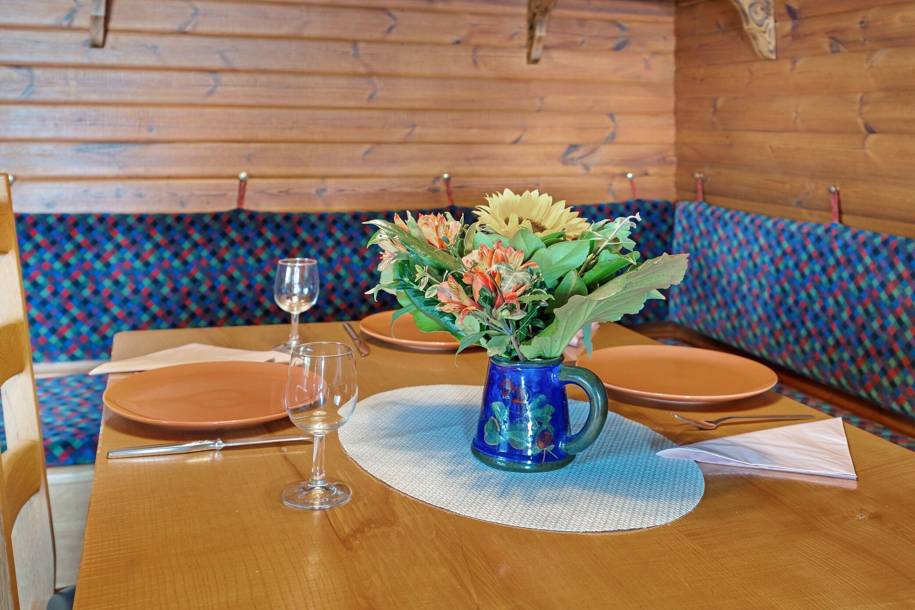 Uma mesa de madeira com um vaso azul de flores, uma taça de vinho e pratos.