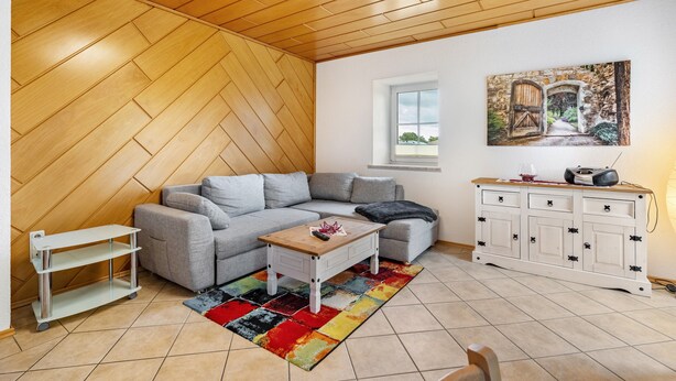 Ein Wohnzimmer mit Fischgrätparkett, einem grauen Ecksofa, einem kleinen Holztisch, einem bunten Teppich, einer weißen Kommode mit Schubladen und einem Gemälde einer rustikalen Tür.