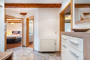 A hallway with a wooden ceiling, a radiator, a bed, and a bathroom with a mirror and sink.