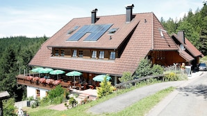 Un edificio de dos pisos con techo inclinado, paneles solares, mesas al aire libre con sombrillas y un coche estacionado cerca.