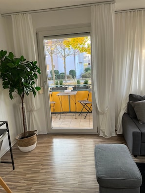 Una sala de estar con un ventanal corredero que da a un espacio exterior con una mesa y sillas, un sofá gris y una planta en maceta.