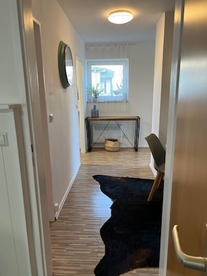 A hallway with a wooden floor, a black rug, a chair, a table with a basket, and a window with curtains.
