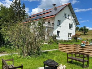 A white building with a red roof, surrounded by greenery and a wooden bench.