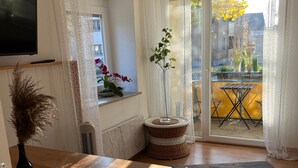 Un salon moderne avec une table en bois, un téléviseur fixé au mur, un tabouret en osier, une plante en pot et une vue sur le balcon extérieur.