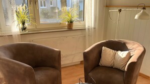 Une pièce avec deux fauteuils marron, une petite table ronde sur laquelle repose un globe terrestre, un tapis en fourrure, une fenêtre ornée de rideaux en dentelle et une plante en pot.