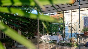 An outdoor patio area with a covered seating arrangement, a table with a bottle and glass, and a backdrop of greenery and a building.