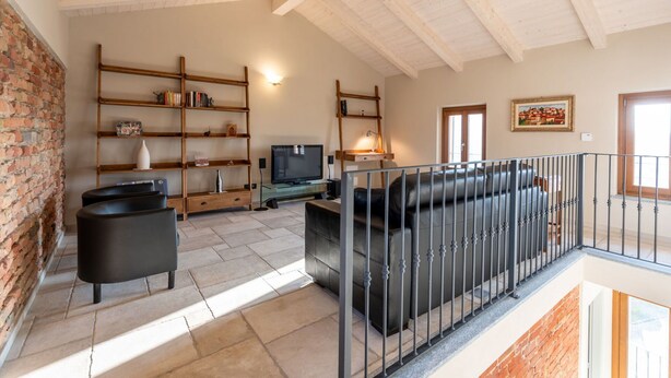 A spacious living area with a brick wall, a wooden ceiling, a black leather sofa, a TV, and a fireplace.