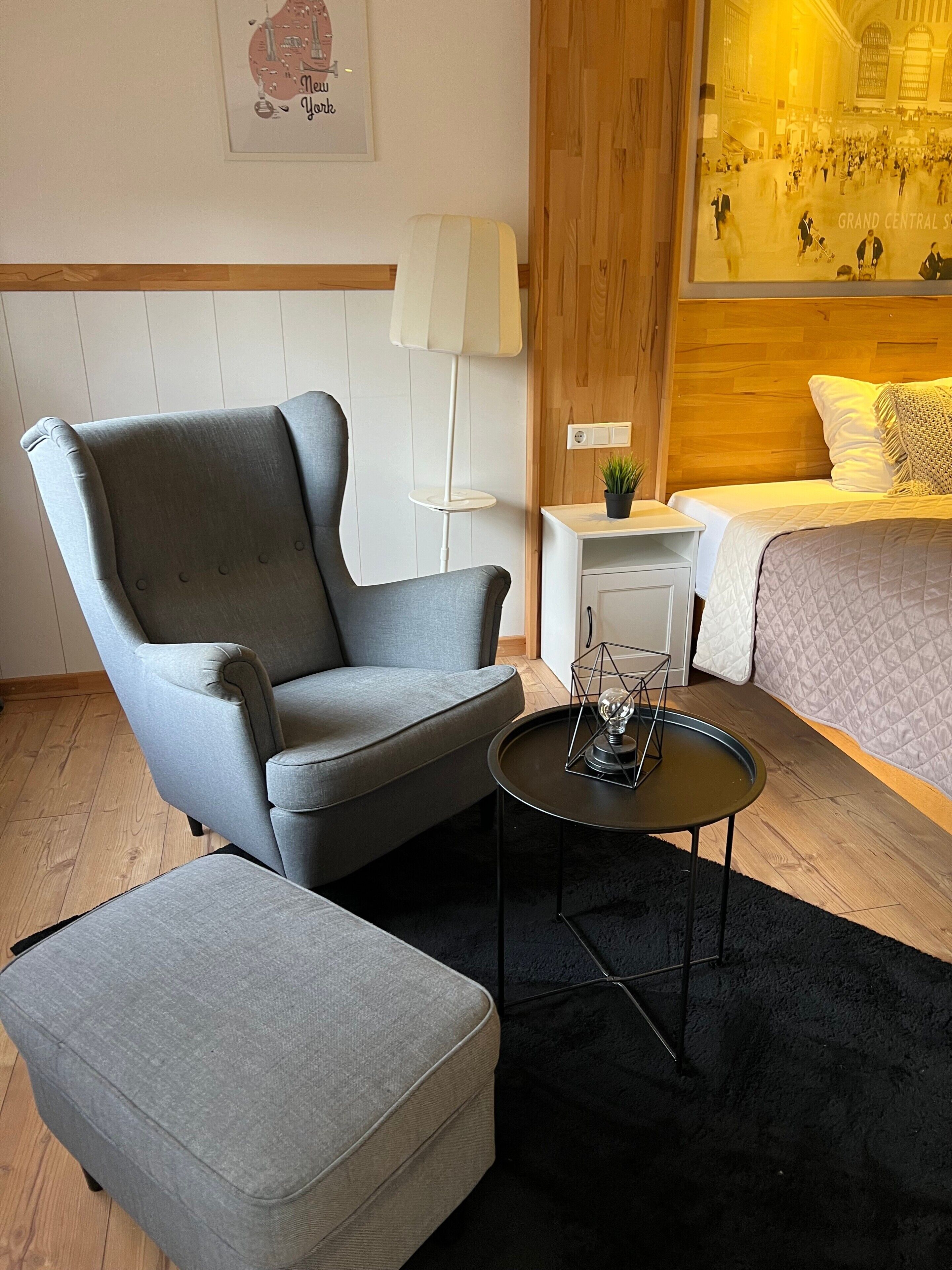 Ein modernes Hotelzimmer mit einem grauen Sessel, einem kleinen runden Tisch mit Lampe, einem Bett mit gesteppter Bettdecke und einem an der Wand befestigten Lichtschalter.