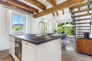 A modern kitchen with a central island, wooden beams, a large window, and a dining area with outdoor seating. - Holiday Home 'Die Alte Mühle Am Hanselishof' with Mountain View, Private Terrace and Wi-Fi (Schenkenzell)
