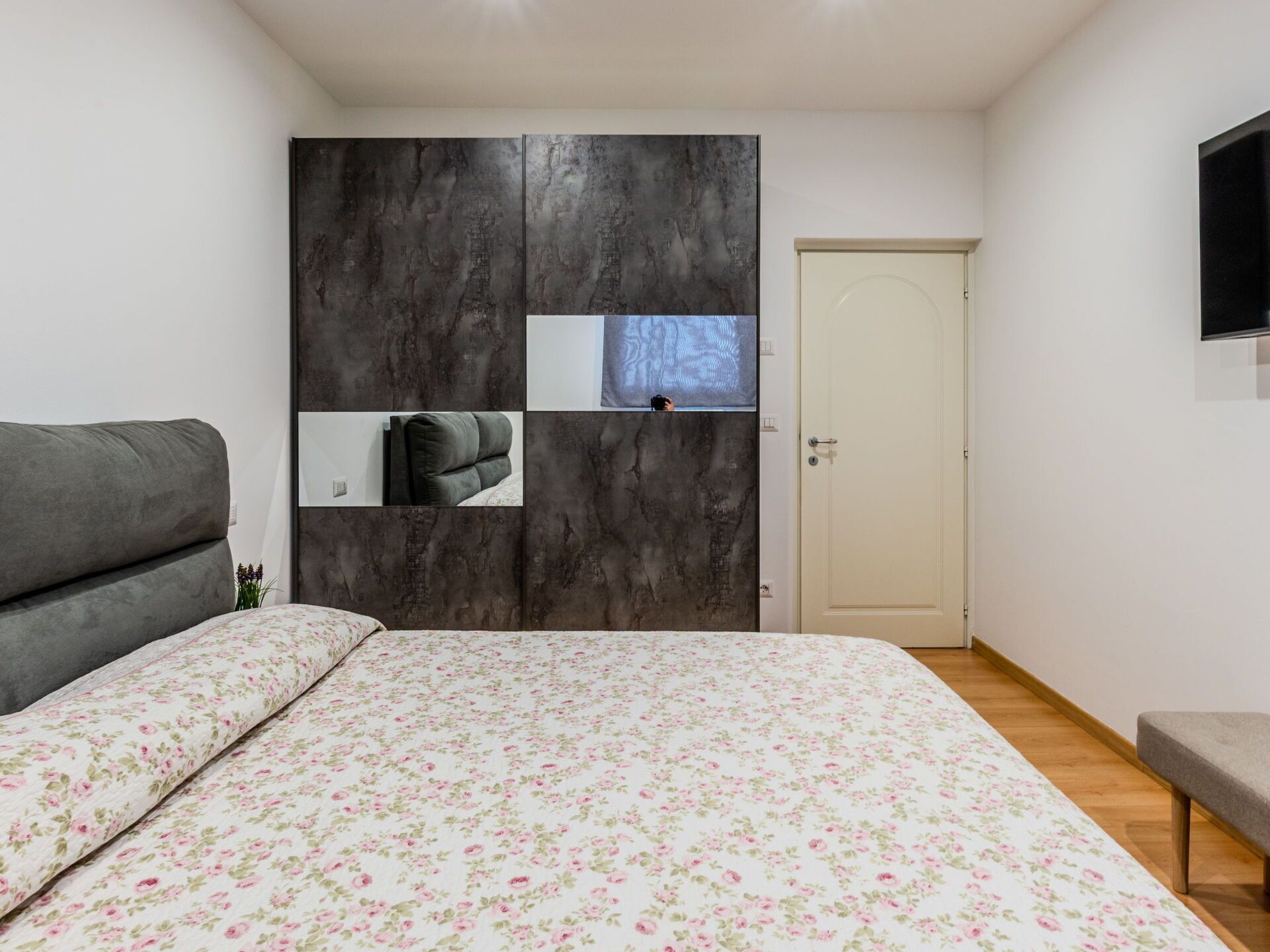 A bedroom with a bed, a grey sofa, a wardrobe with mirrors, and a television on the wall.