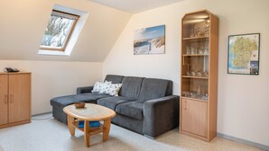 Ein gemütliches Wohnzimmer mit grauem Sofa, einem Holzschrank, einem kleinen runden Tisch, einem Oberlicht und einer gerahmten Karte an der Wand.