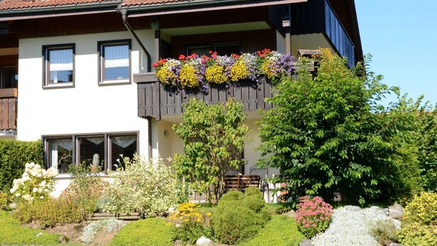 Una casa con techo inclinado, paneles solares, un balcón con jardineras y un jardín bien cuidado.