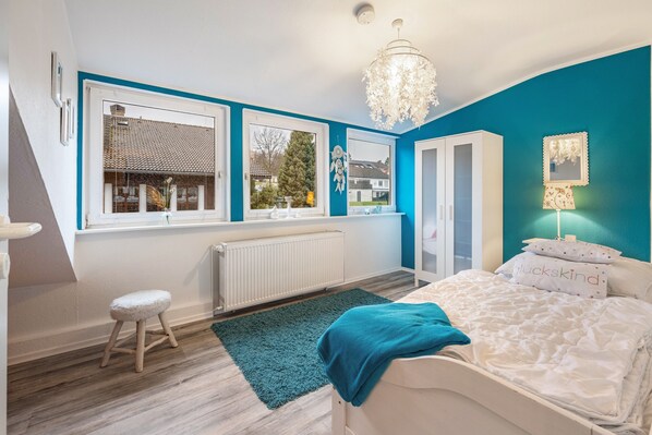 A bedroom with a bed, a window, a radiator, a stool, and a chandelier.