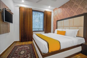 Una habitación de hotel con una cama grande, un televisor montado en la pared, un ventanal con cortinas y una alfombra estampada.