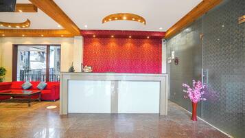 Zona de recepción con pared de diseño rojo, mostrador de recepción, sofá rojo y un jarrón de flores.