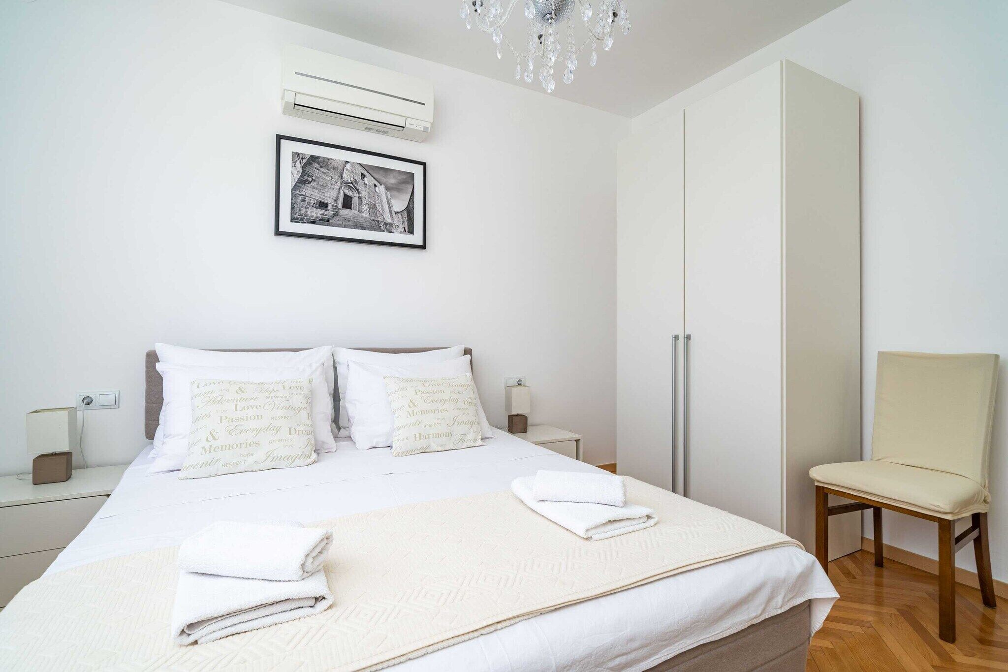 Une chambre à coucher bien rangée, avec un lit, des oreillers, une table de chevet, une chaise, un climatiseur fixé au mur, un lustre et un tableau encadré.