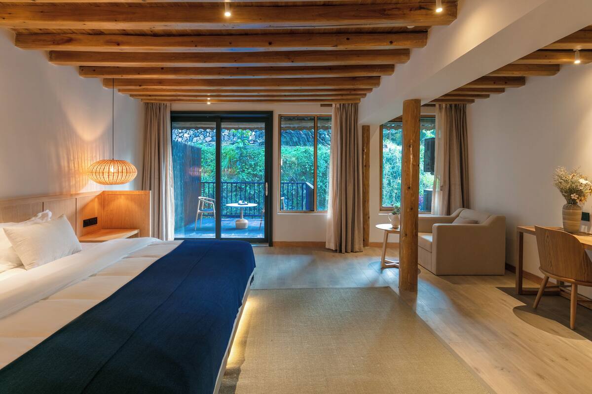 A modern hotel room with a wooden ceiling, a large bed, a desk, and a view of a lush garden through the windows.