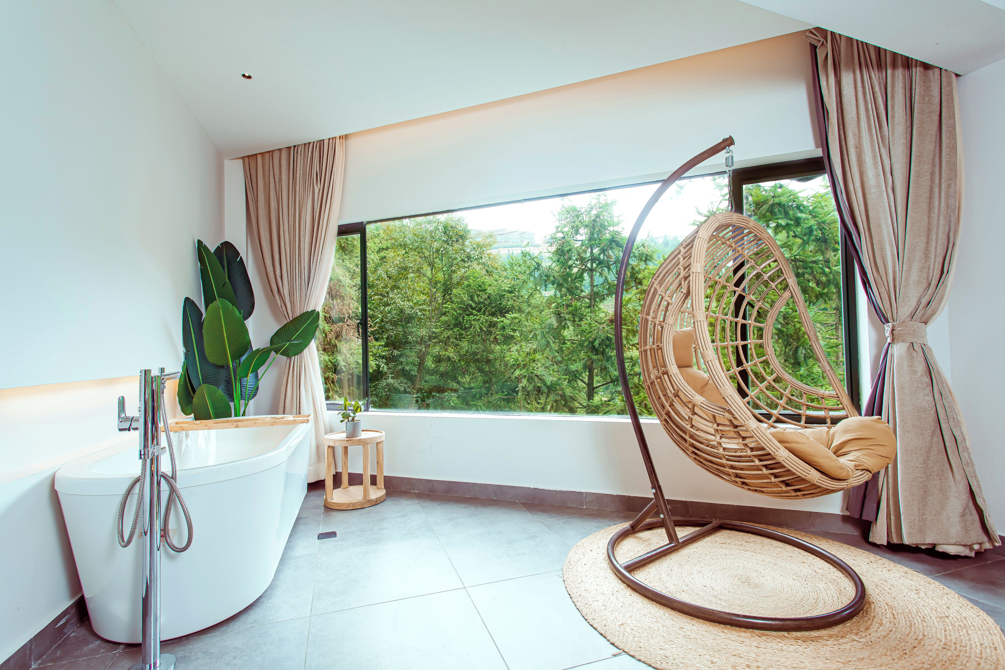 A modern bathroom with a freestanding bathtub, a hanging chair, and a large window offering a view of greenery.