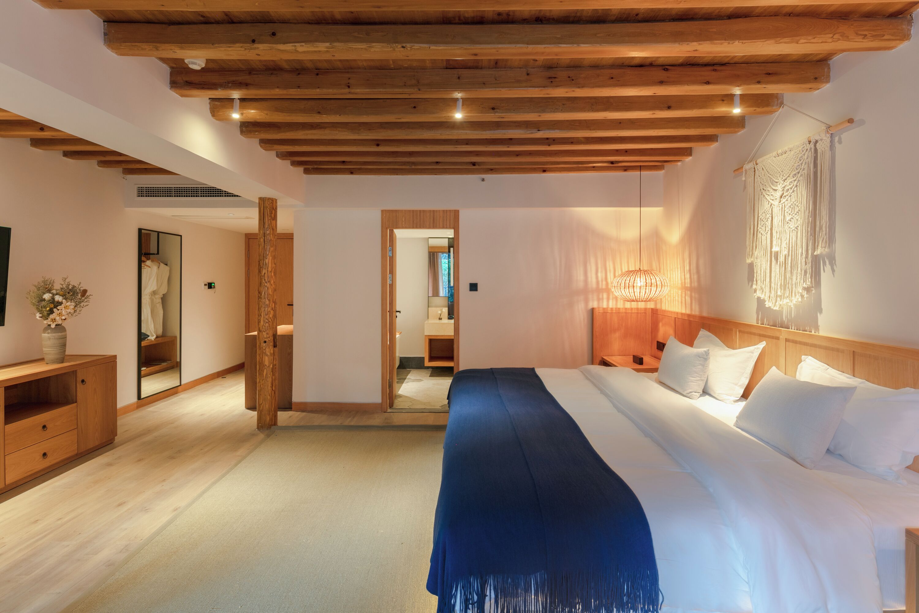 A bedroom with a large bed, wooden ceiling beams, a mirror, a bedside table, and a macrame wall hanging.