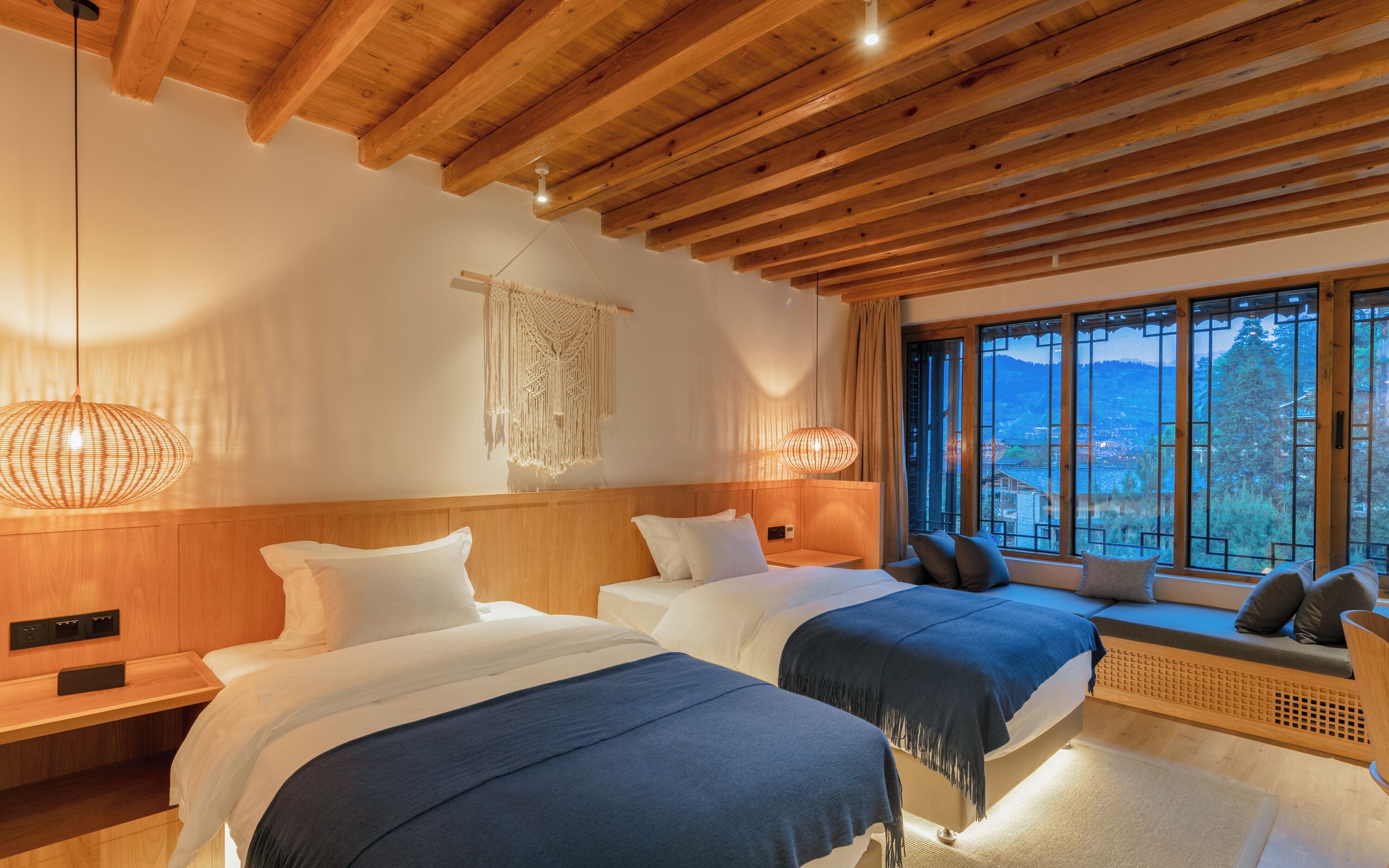 Une chambre confortable avec deux lits, des poutres apparentes en bois au plafond, des décorations murales en macramé et une grande fenêtre offrant une vue panoramique.