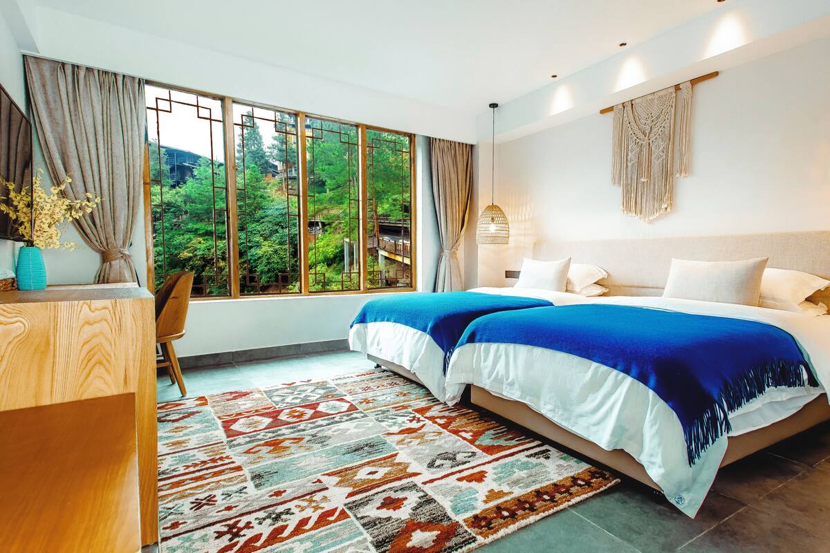 A modern bedroom with a large bed, a desk, and a view of greenery outside.