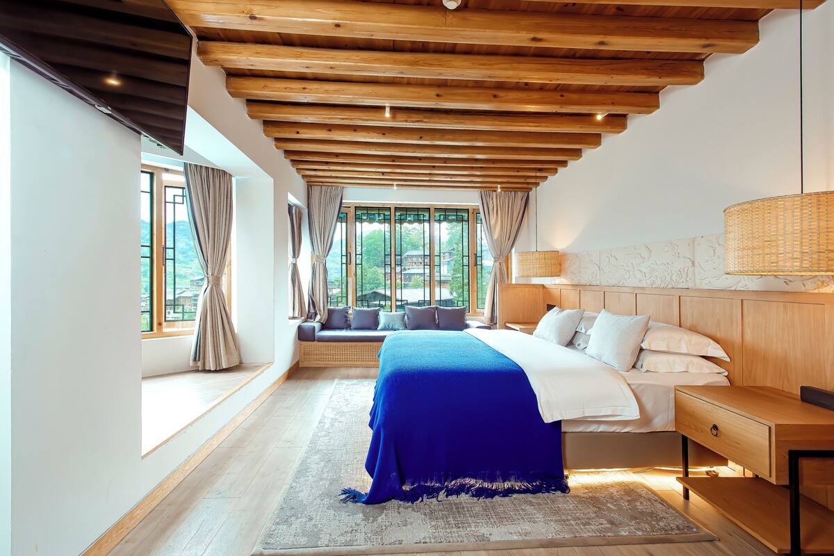 A bedroom with a large bed, wooden ceiling beams, a window seat, and a view of greenery.