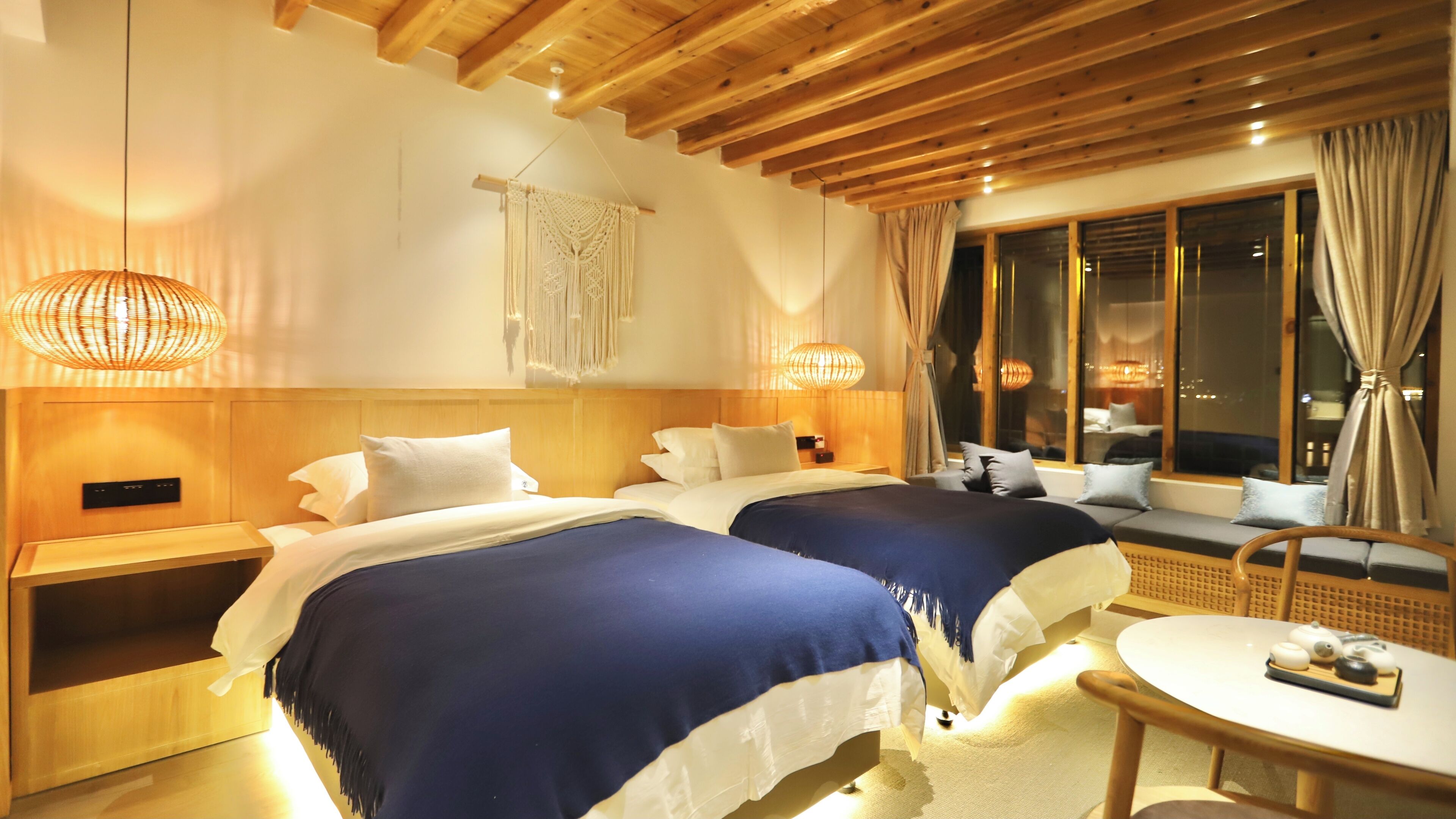 Una camera d'albergo con due letti, poggiatesta in legno, un'ampia finestra con tende e un piccolo tavolino rotondo con teiera.