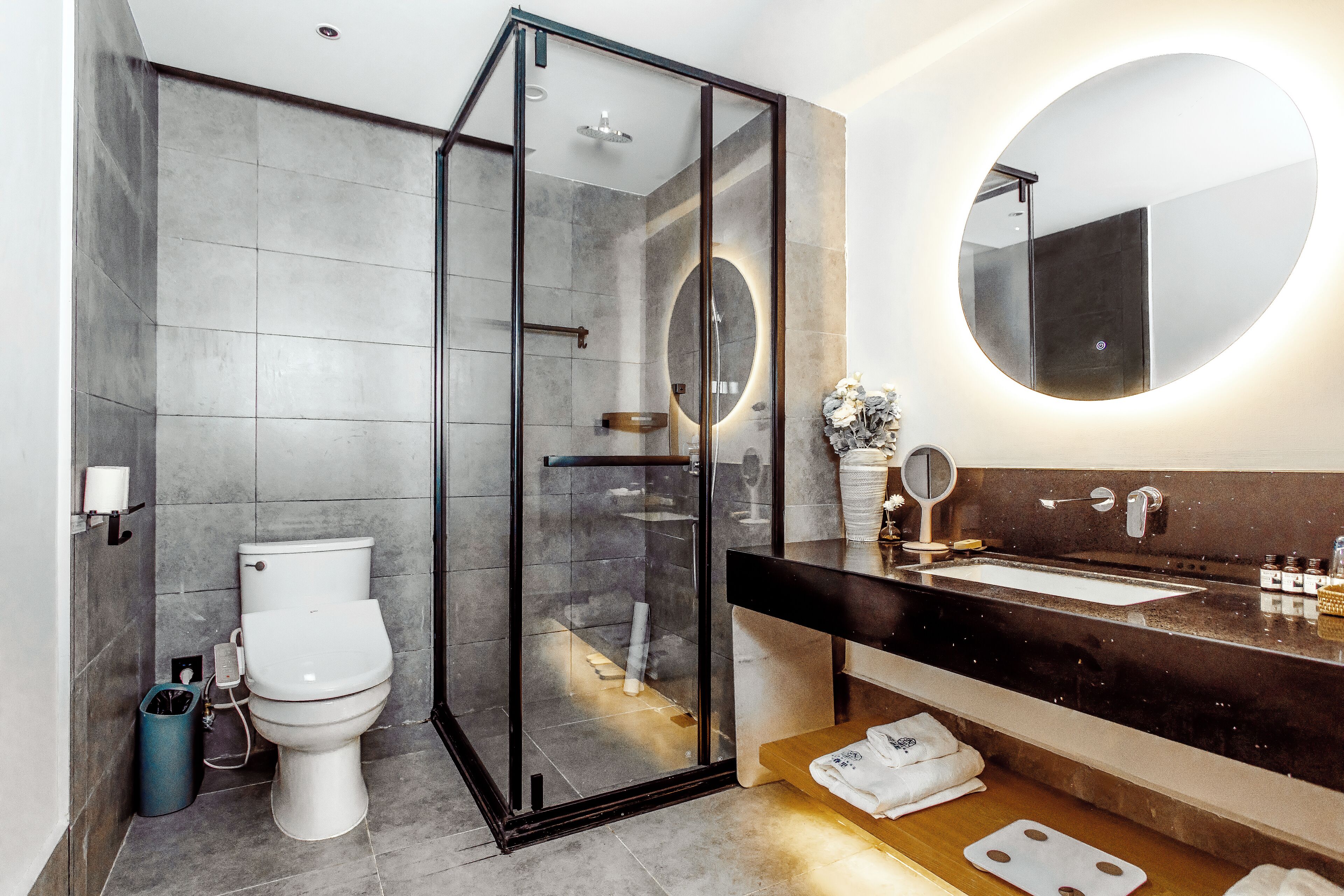 Ein modernes Badezimmer mit einer einteiligen Duschkabine, einer weißen Toilette, einem dunklen Waschtisch und einem runden Spiegel.