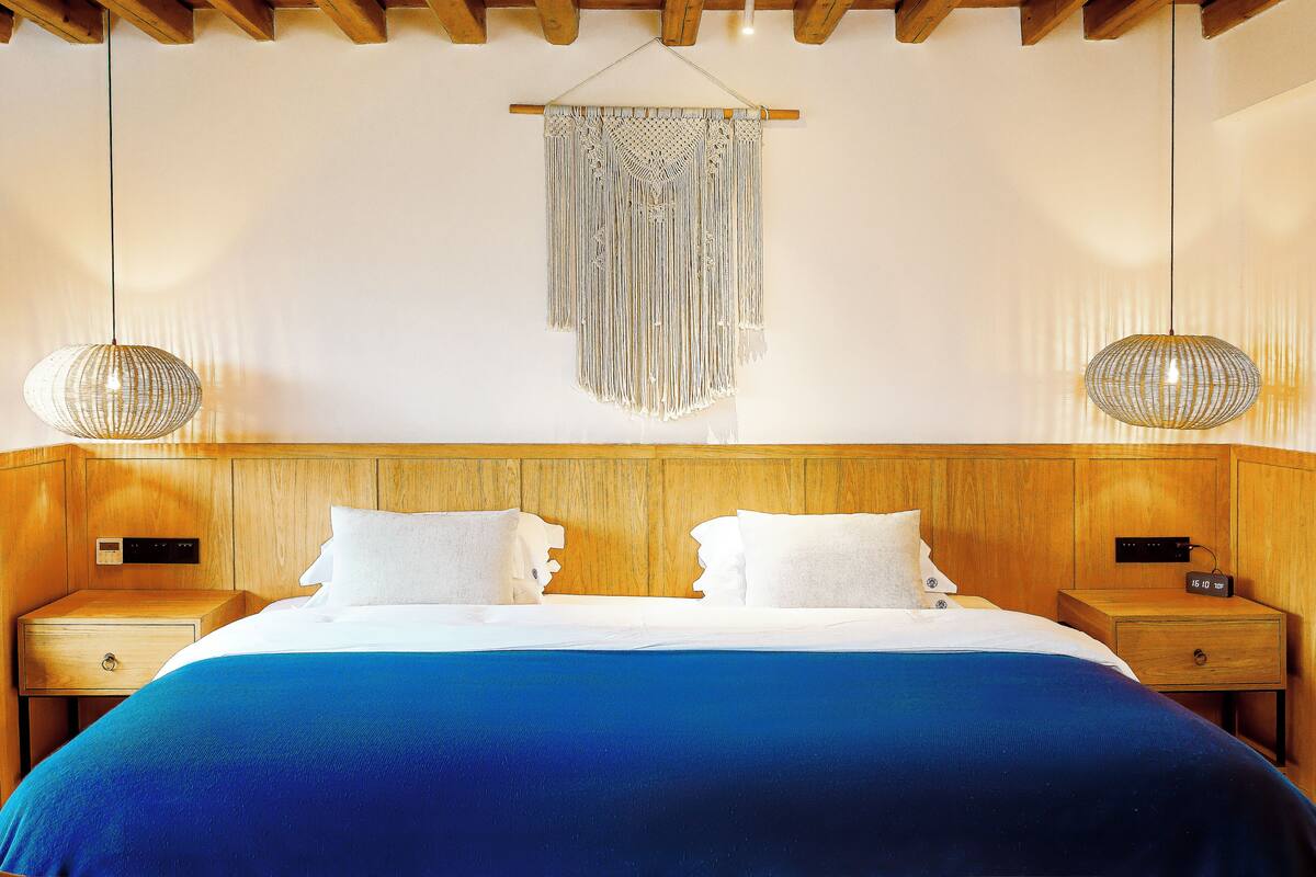 A hotel room with a wooden headboard, a large bed with blue bedding, two bedside tables with lamps, and a macrame wall hanging.