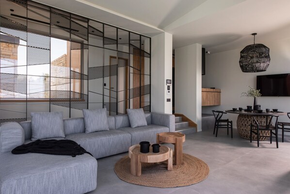 A modern living room with a grey sectional sofa, a round wooden coffee table, and a dining area with a round table and chairs. - EGO Villas in Vasiliki (Lefkada)