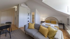 Habitación de hotel moderna con techo estilo loft, sofá gris con cojines amarillos, cama con cabecera de madera y una pequeña mesa con una silla.