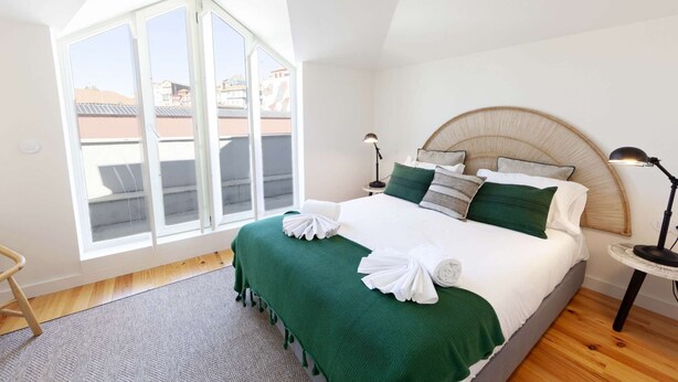 A bedroom with a large window, a bed with green bedding, and a bedside table with a lamp.