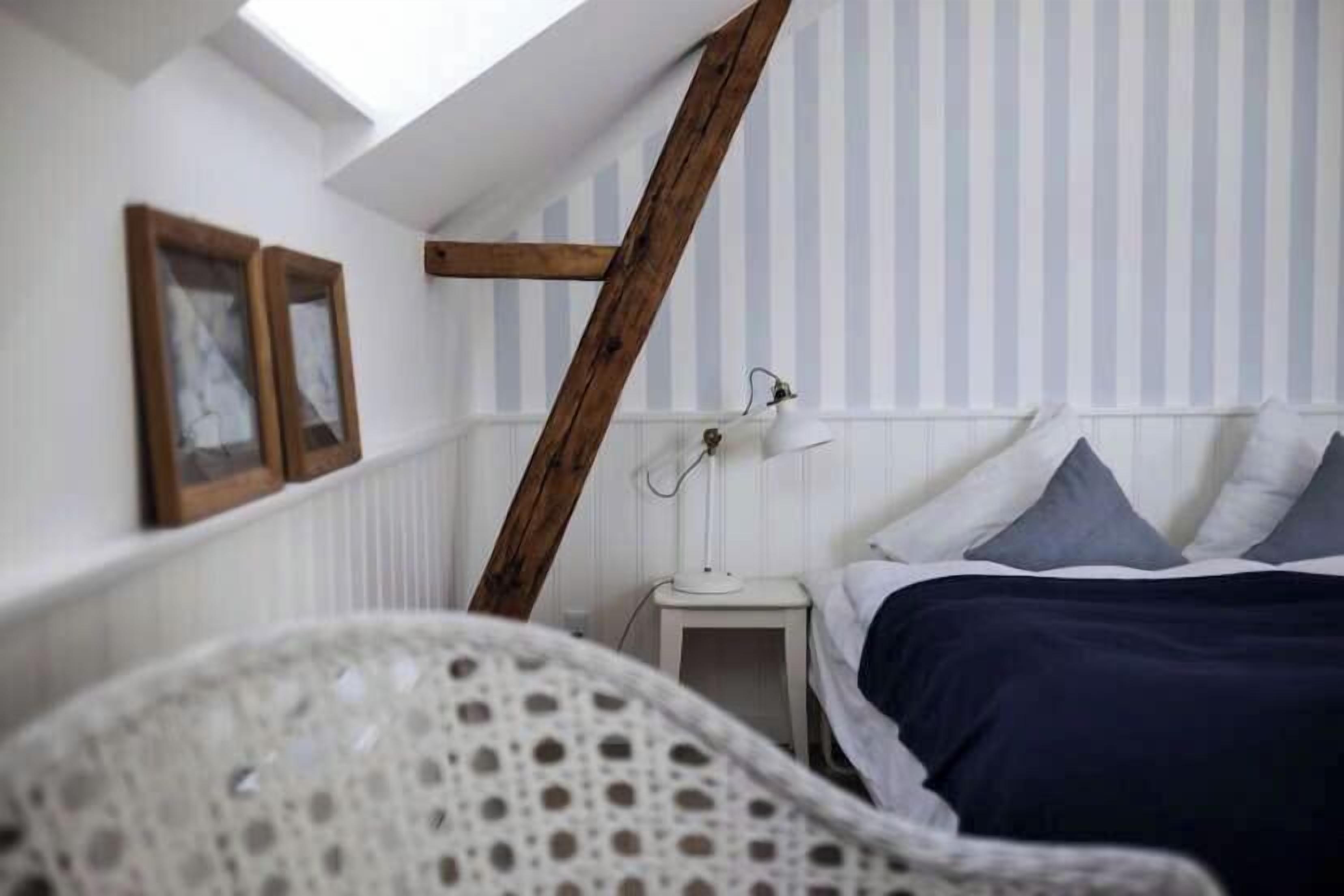 A bedroom with a sloped ceiling, a bed with dark bedding, a small bedside table with a lamp, and a chair.